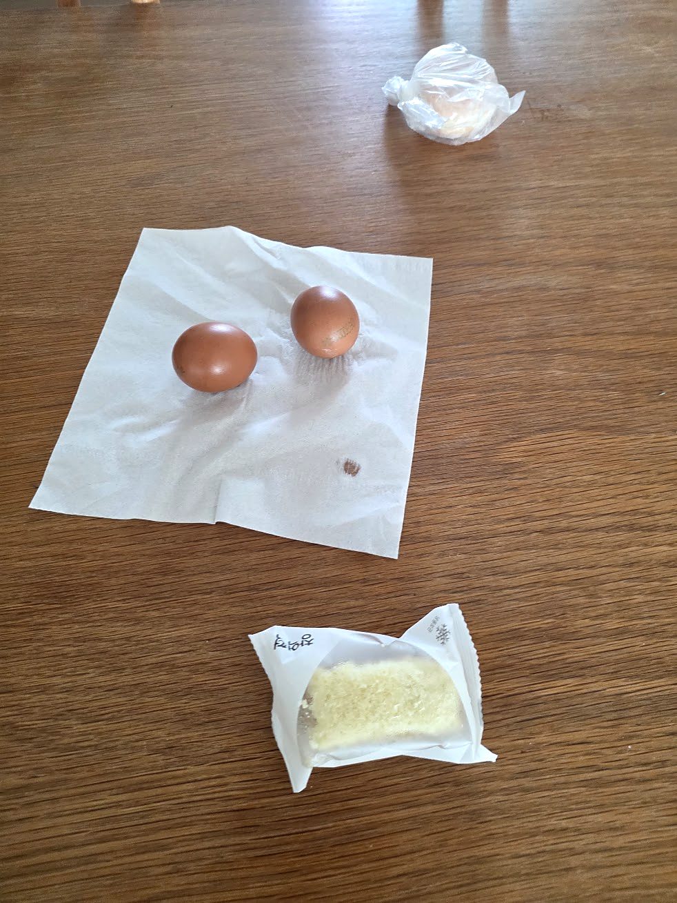 A Korean kid's breakfast: two boiled eggs on a napkin, a packaged rice cake and a doughnut wrapped in plastic on a wooden table